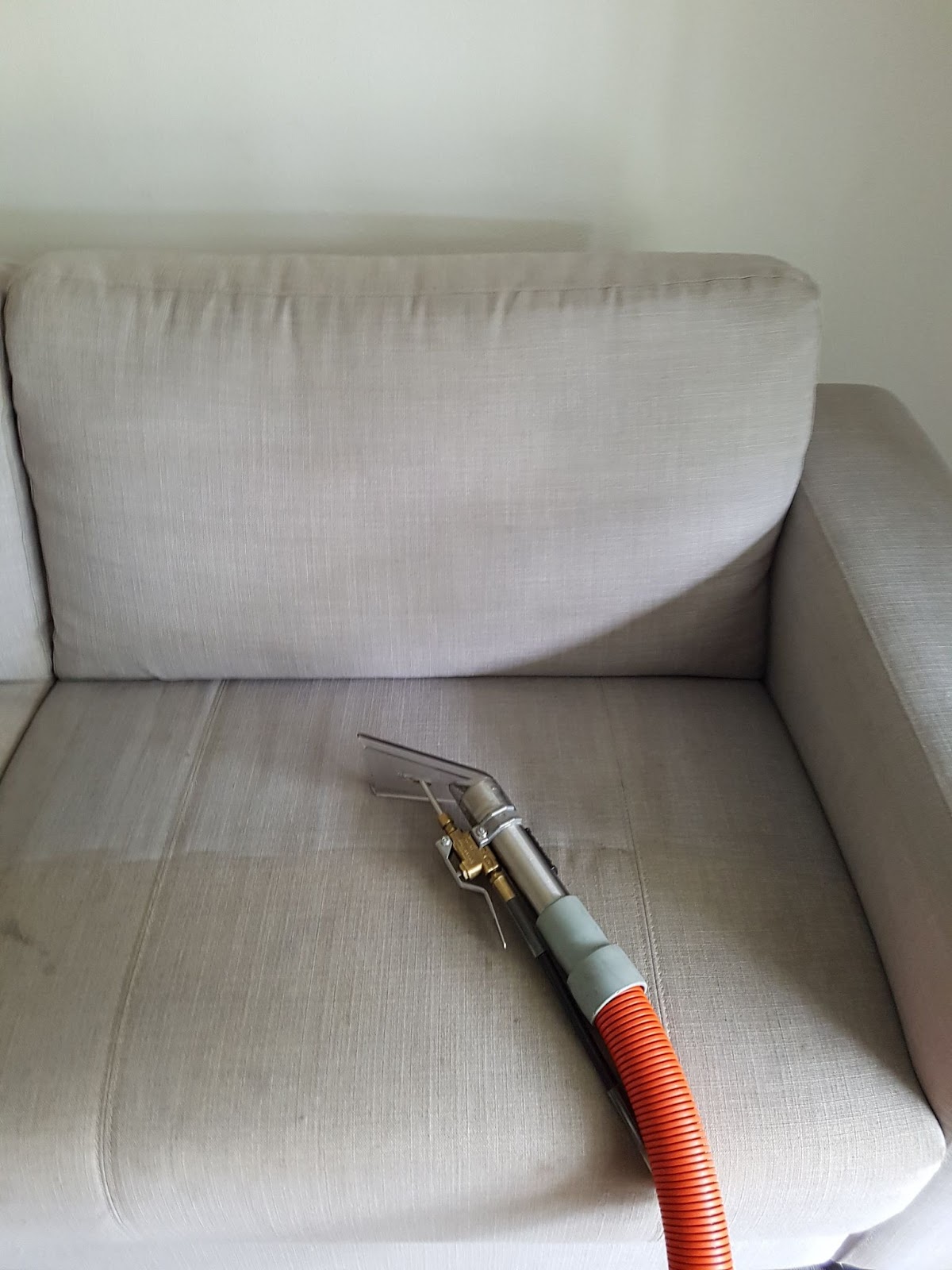 A light grey fabric armchair having its seat cleaned with an upholstery tool on an orange hose